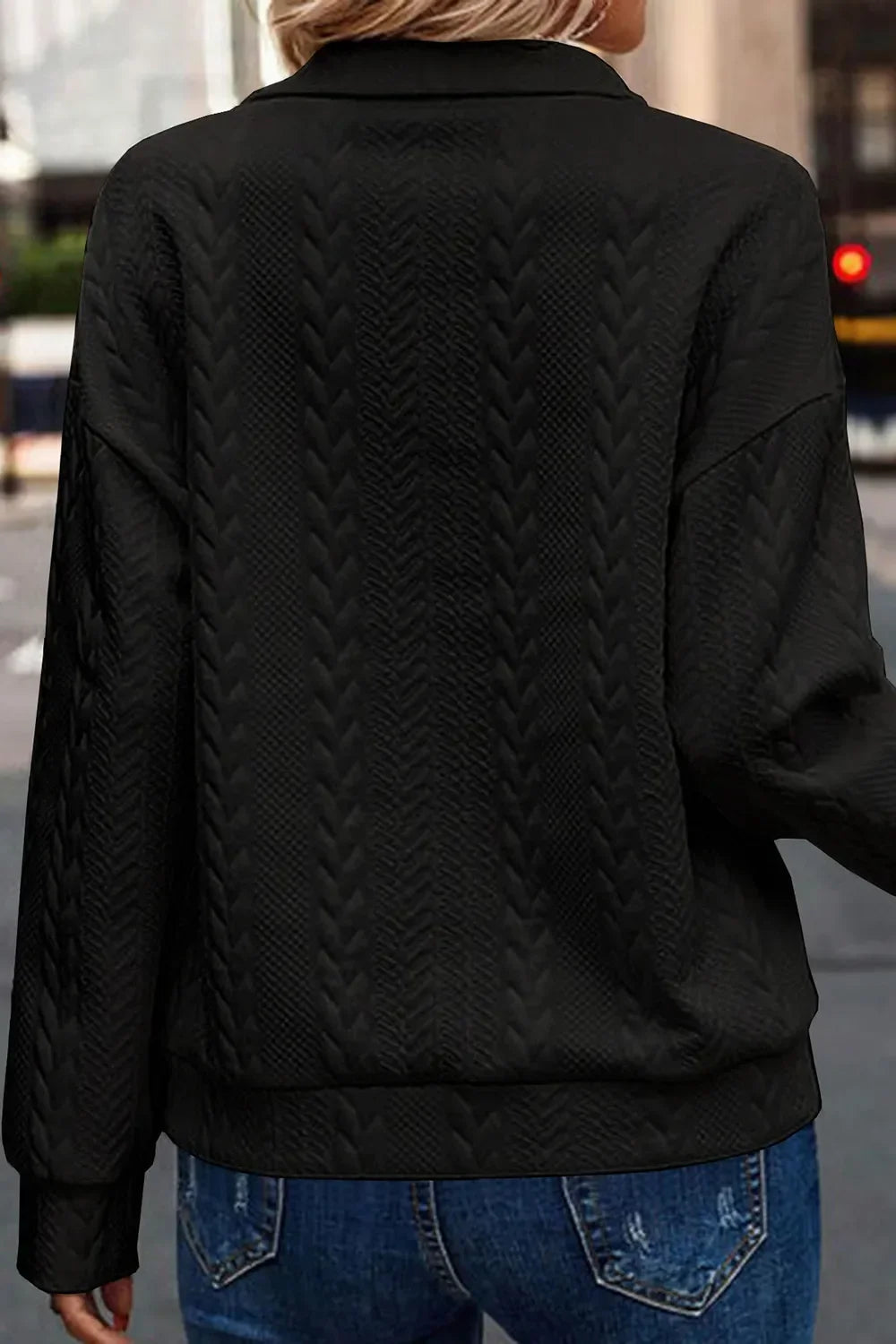 Back view of a woman wearing a textured black sweater with chevron knit pattern and blue jeans.
