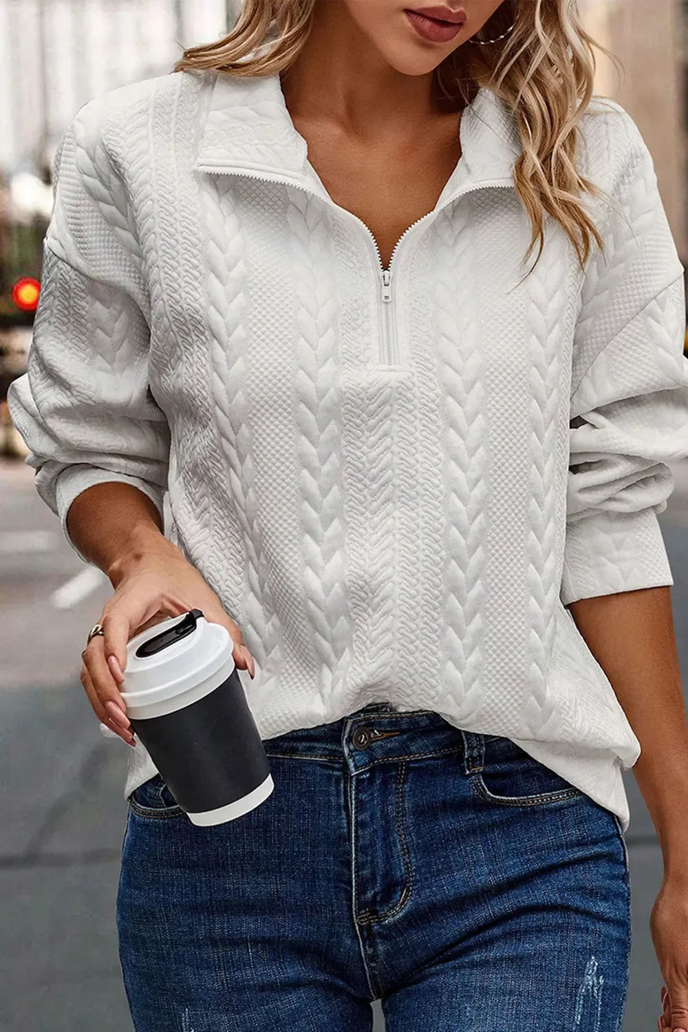Woman in textured white zip-up sweater and blue jeans holding coffee cup on city street